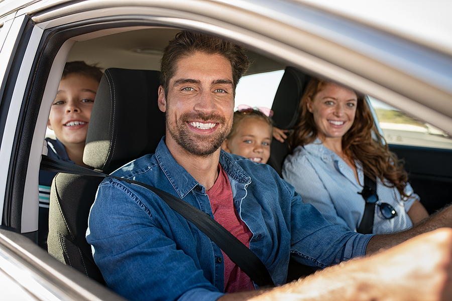 Smiling young family with two children sitting in car and driving. Family relaxing during road trip while looking at camera. Portrait of happy father riding in a car with wife, son and daughter.